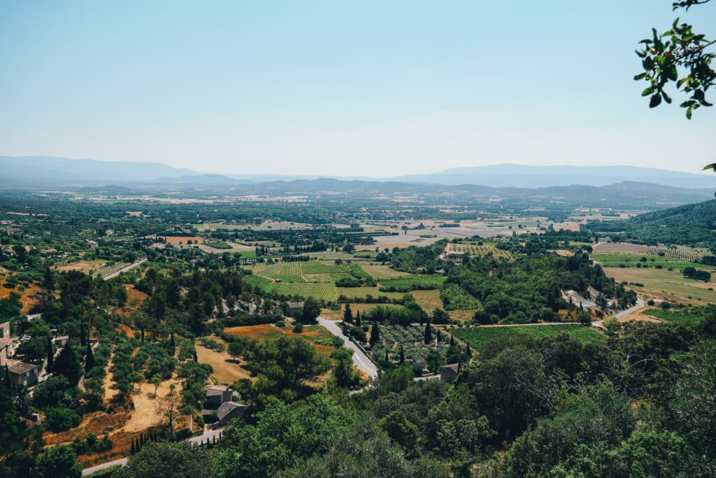Luberon countryside panorama near Gordes, showing patchwork fields, rolling hills, and distant mountains under a blue sky.