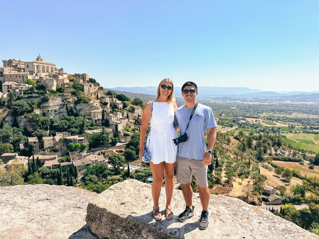 Two travelers posing at a Gordes viewpoint with the stone village perched on a rocky hill in the background.