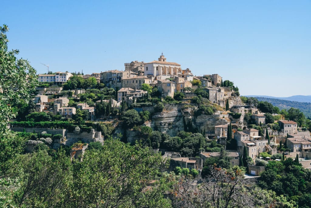 Wide panoramic view of Gordes village in Provence, with stacked stone houses and greenery on the hillside.