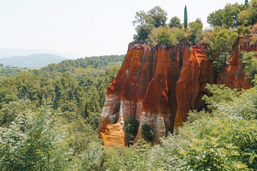 Close view of the ochre cliffs in Roussillon with textured red and yellow rock faces and pine trees above.