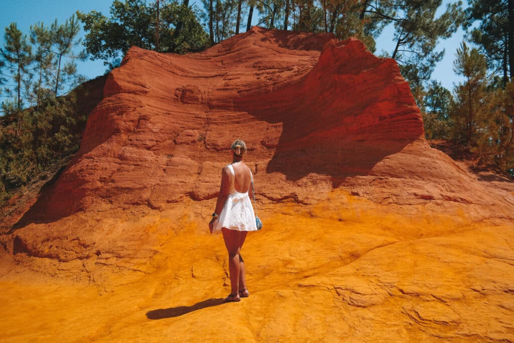 Hiker walking through towering ochre cliffs on Le Sentier des Ocres trail in Roussillon, showing the scale of the canyon.