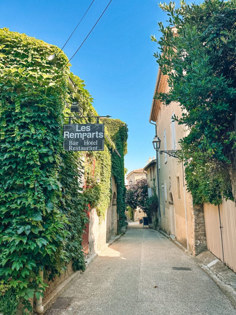 Narrow sunlit lane in Venasque by the Hotel les Ramparts, lined with tall green hedges and pale stone buildings under a bright blue sky.