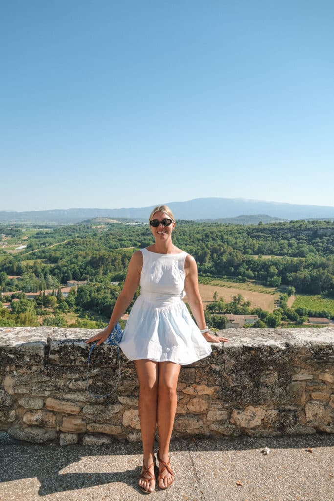 Woman in a white dress standing at a stone viewpoint wall with sweeping Provençal countryside views.