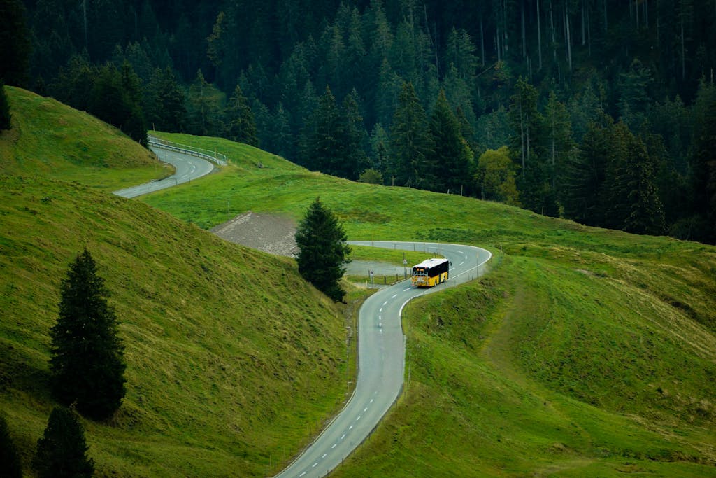 A yellow bus travels along a winding road through lush green hills and forests in Switzerland.