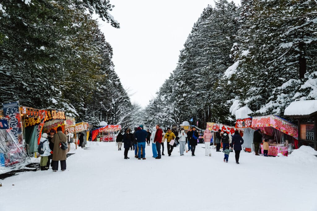 Winter food stalls lining the snowy approach to Hokkaido Jingu with crowds walking through.