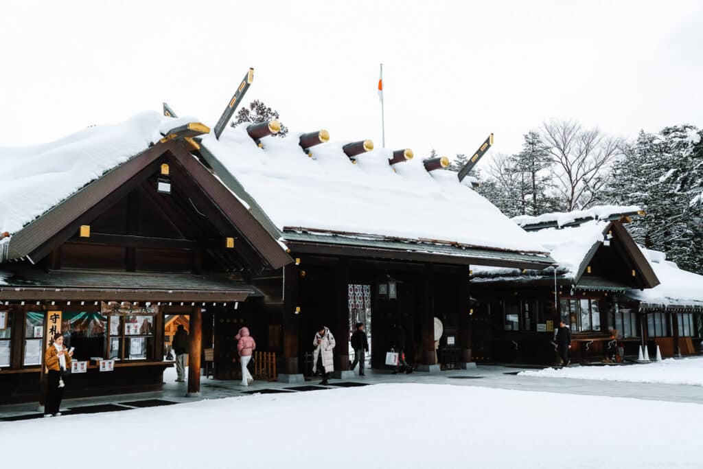 Wide view of Hokkaido Jingu buildings with snow on the roofs and visitors gathered near the entrance.