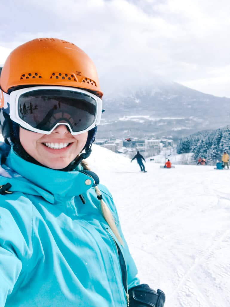Smiling skiier in teal jacket and goggles posing for a close-up portrait on a snowy slope in front of Mount Yotei