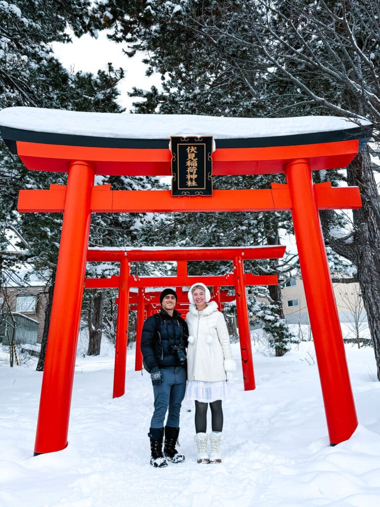 Two visitors standing beneath the bright red torii entrance at Sapporo Fushimi Inari Shrine, surrounded by snow-covered trees.