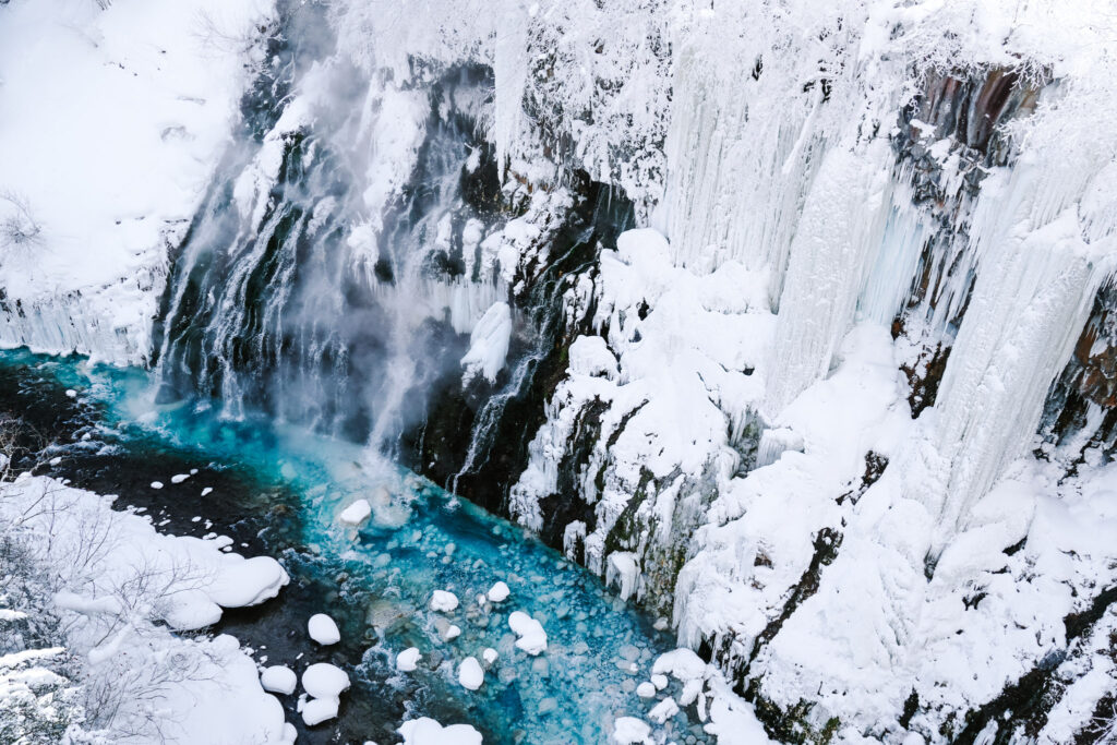 Close wide shot of Shirahige Waterfall cascading into vivid blue water with snow and ice along the banks, Hokkaido