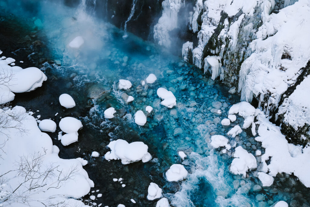 Steaming blue river below Shirahige Waterfall with snow-covered boulders in Shirogane Onsen, Hokkaido.