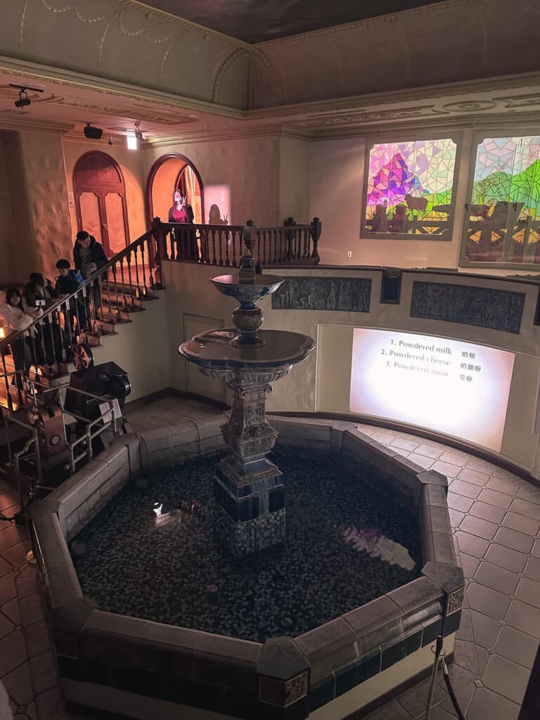 Dim CHOCOTOPIA room with a central tiered fountain, balcony staircase, and warm European-style decor.