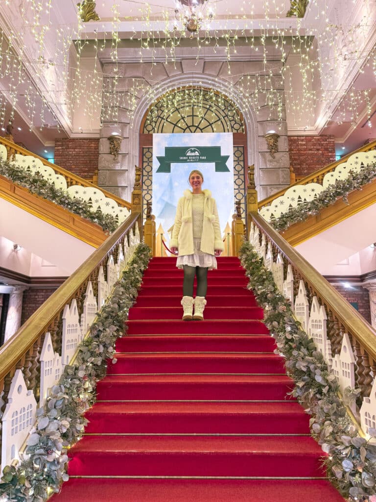 Ornate red-carpeted staircase lined with garlands and fairy lights inside the CHOCOTOPIA mansion.