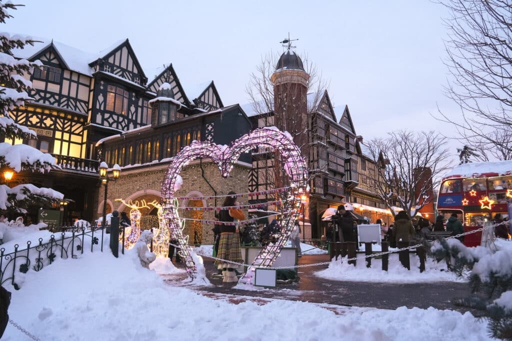 Heart-shaped light arch surrounded by snow, with storybook-style buildings glowing behind during the winter illumination.