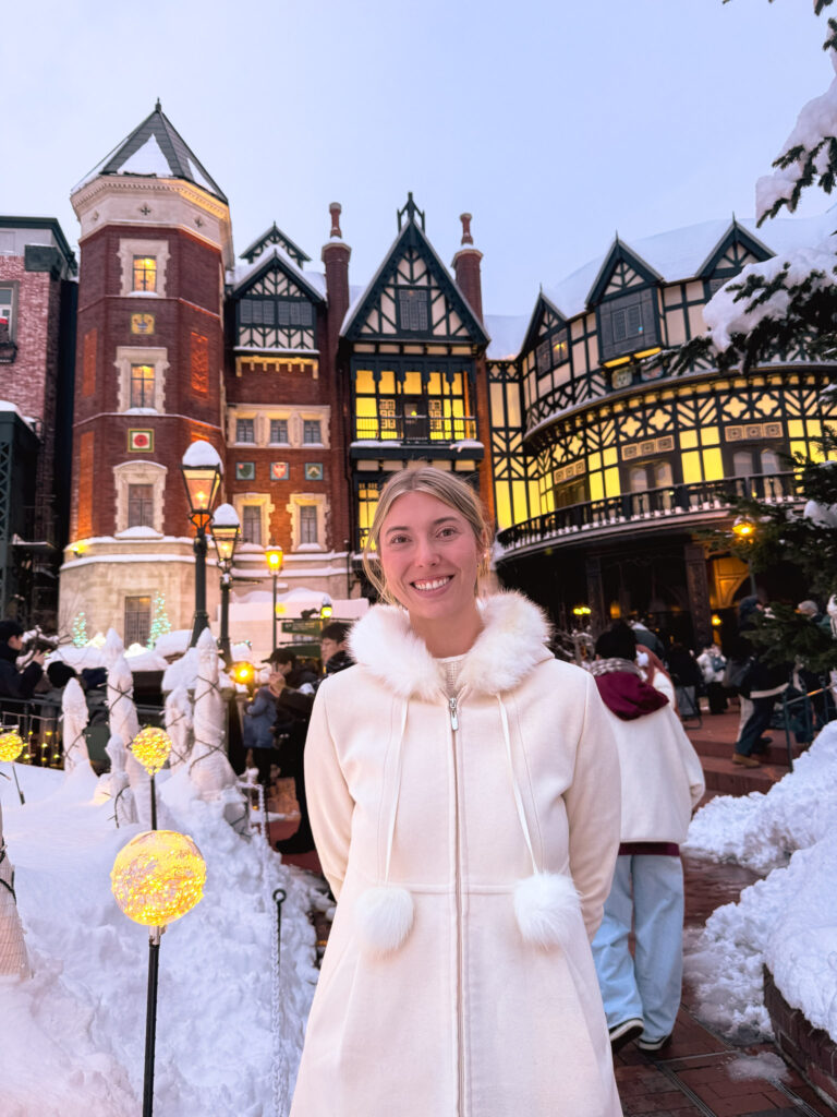 Person in a white coat smiling in a snowy outdoor courtyard, surrounded by warm lights and European-style buildings at night.