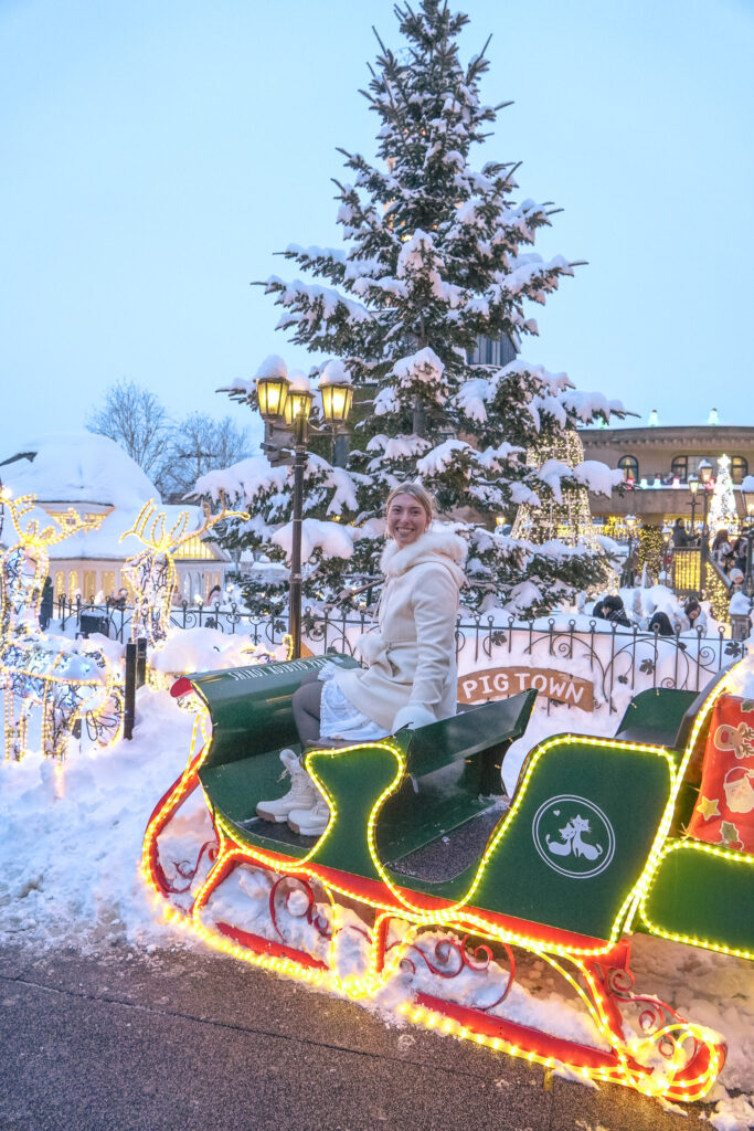 Green sleigh outlined with colorful lights beside a snow-covered tree and glowing buildings during Shiroi Koibito Park’s winter illumination.
