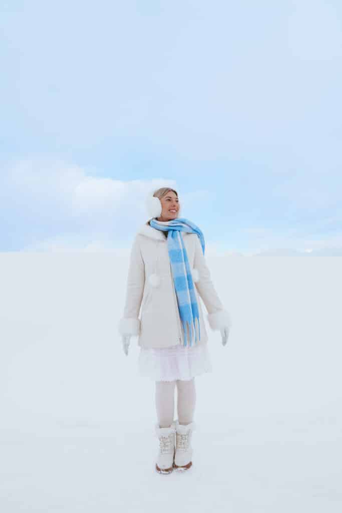 A person in a white coat and light blue scarf stands amid Biei's gently rolling snow-covered hills, with the iconic lone Christmas Tree visible in the soft white background.