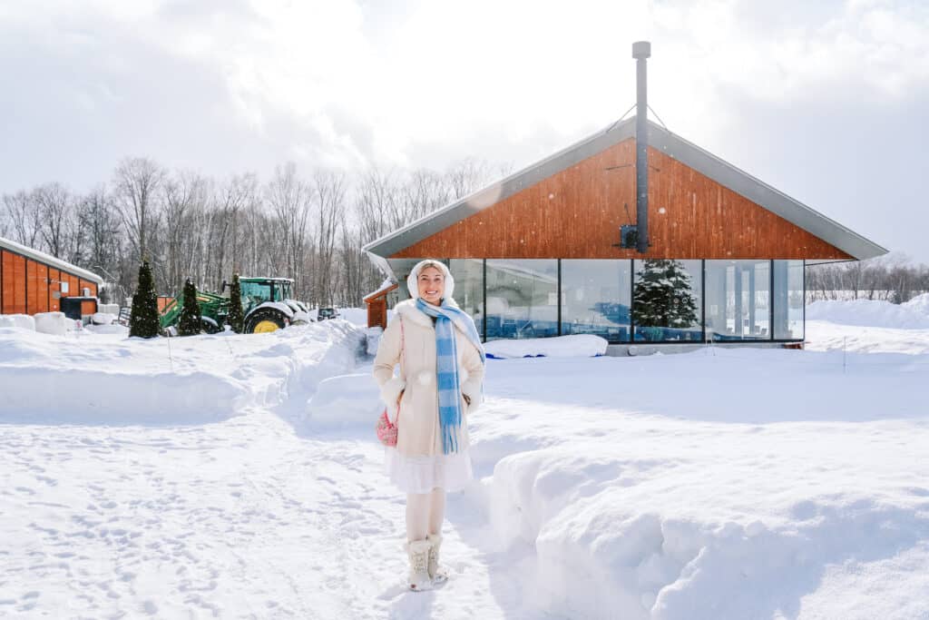 Woman in white standing in the snow in front of the modern farm buildings at La Laiterie Ferme Biei