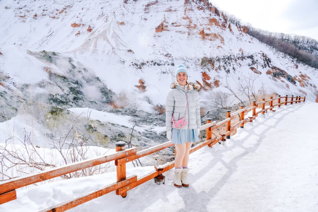 A traveler in front of a snowy view of Noboribetsu Jigokudani Hell Valley with boardwalk trail and steaming volcanic landscape in Hokkaido, Japan