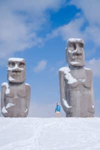 A visitor in a very cute outfit standing next to Moai statues towering above snowy landscape at Makomanai Takino Cemetery near Sapporo in winter