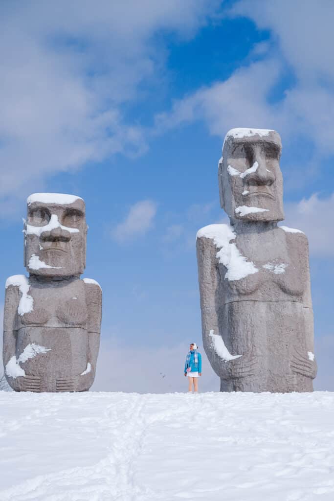 A visitor in a very cute outfit standing next to Moai statues towering above snowy landscape at Makomanai Takino Cemetery near Sapporo in winter