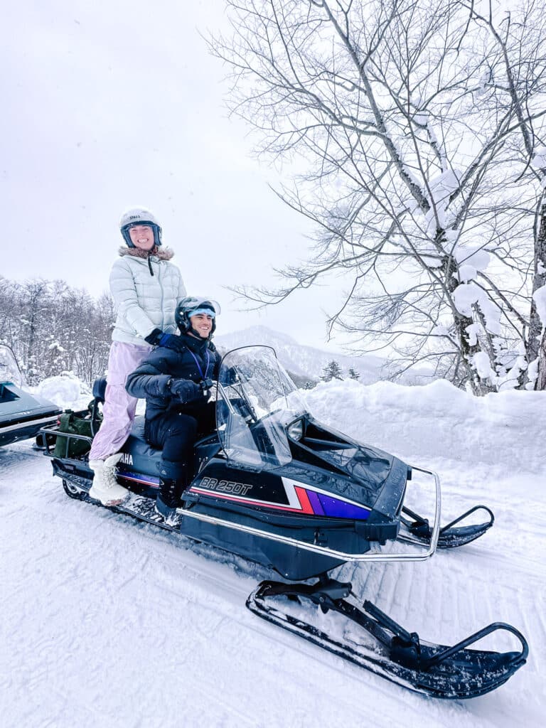 Two smiling riders sitting on a snowmobile during a guided snowmobiling tour at Snow World Toya in Hokkaido