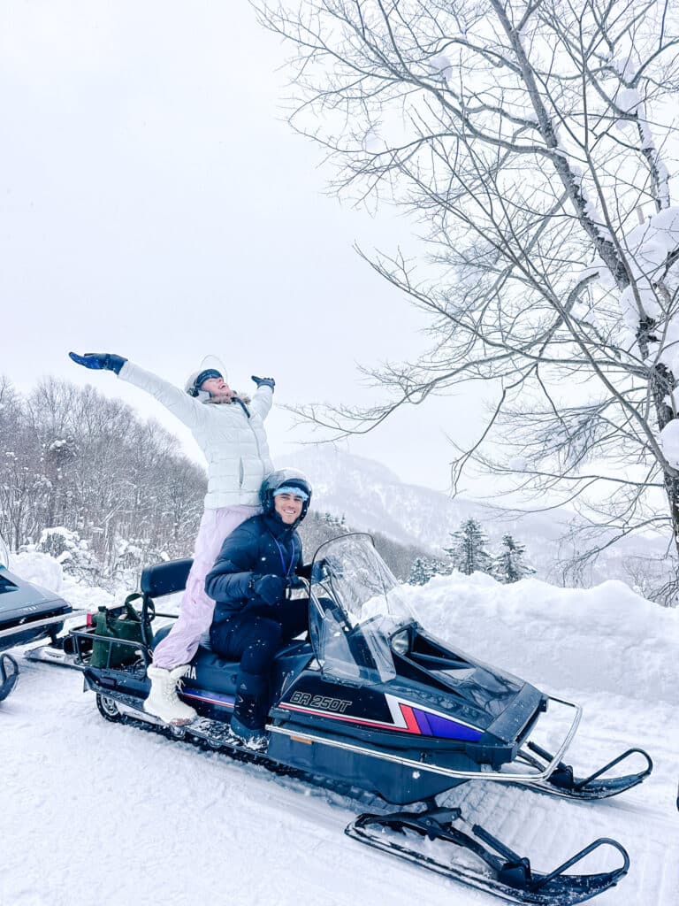 Snowmobiler celebrating with by standing on a snowmobile with raised arms besides a scenic lookout in Hokkaido