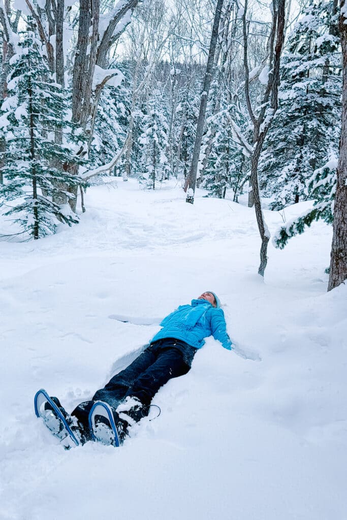 A snowshoer in a bright blue puffer in the Shirogane forest making a snow angel in snow drift