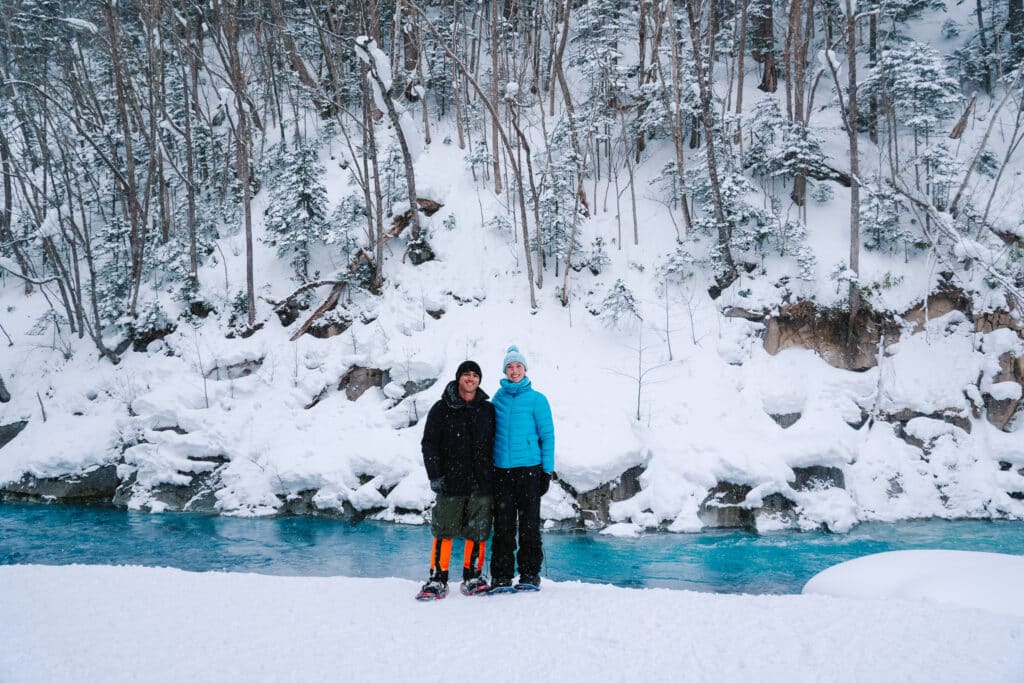Couple standing beside the Shirogane Blue River during a snowshoeing tour in Biei, Hokkaido