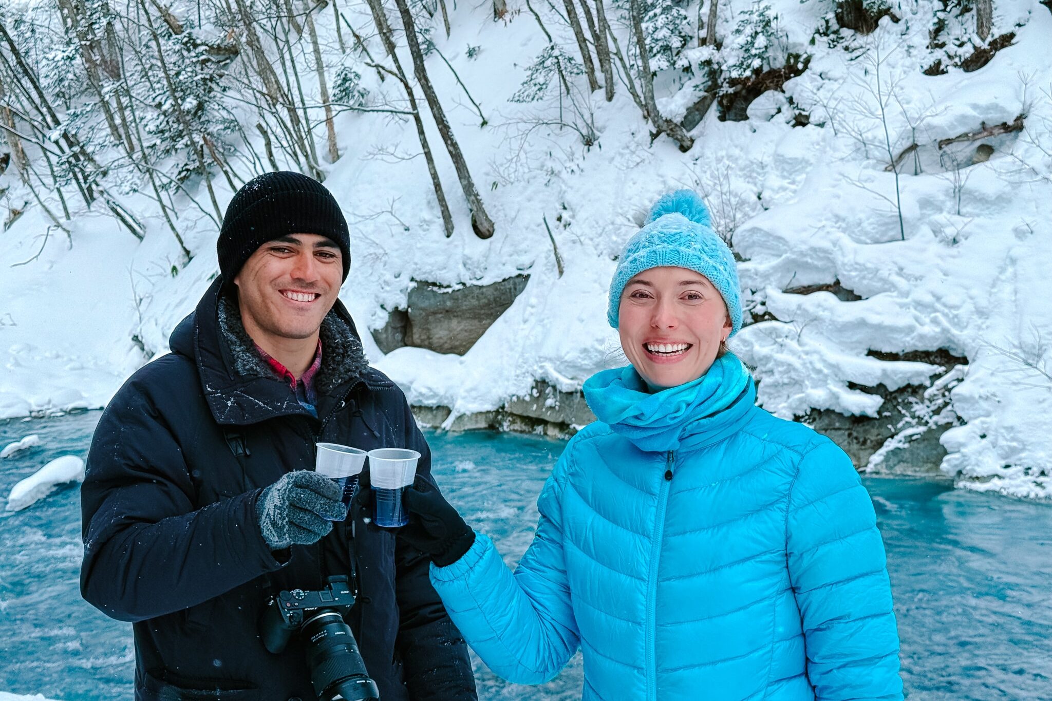 Two snowshoers toasting with steaming cups of tea beside the bright blue Shirogane River during a winter adventure in Biei