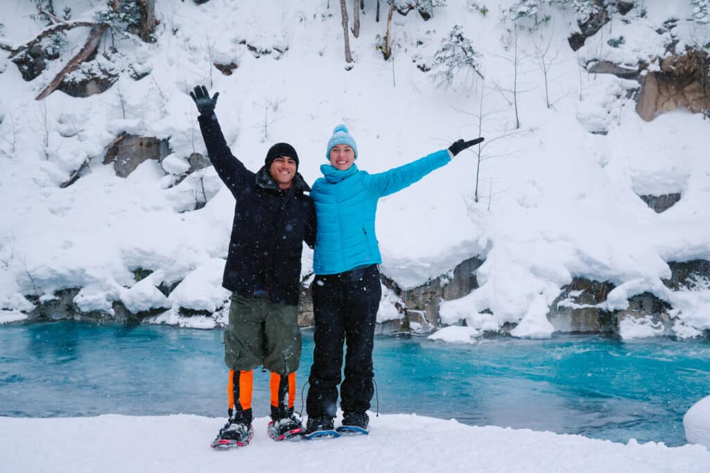Two snowshoers celebrating beside the bright blue Shirogane River during a winter adventure in Biei