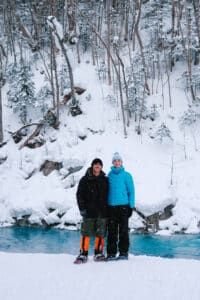 Couple standing beside the Shirogane Blue River during a snowshoeing tour in Biei, Hokkaido