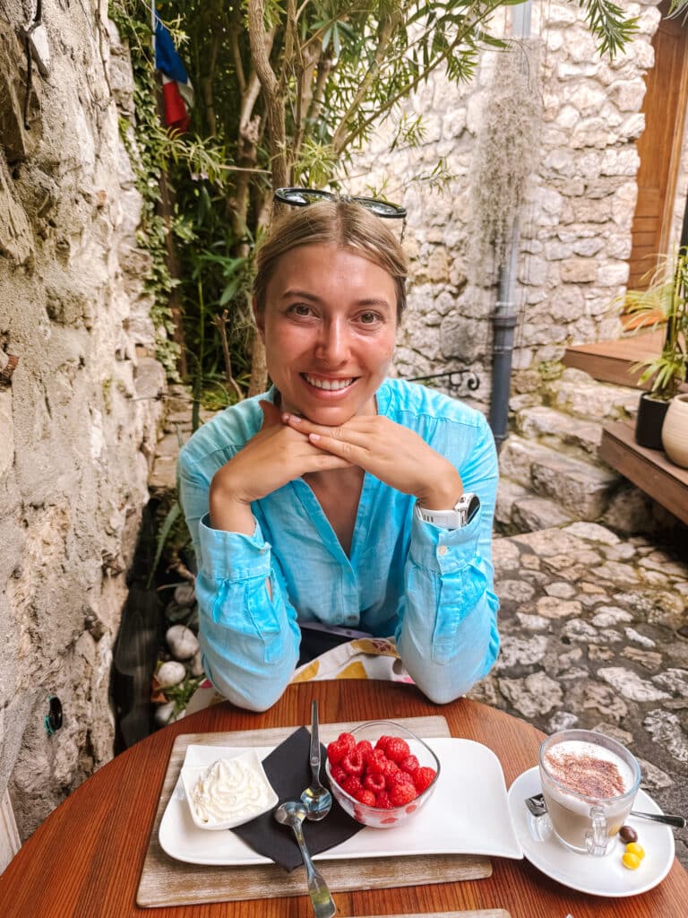 Woman sitting at a café table at Restaurant Deli in the medieval village of Eze on the French Riviera, with stone walls and greenery in the background
