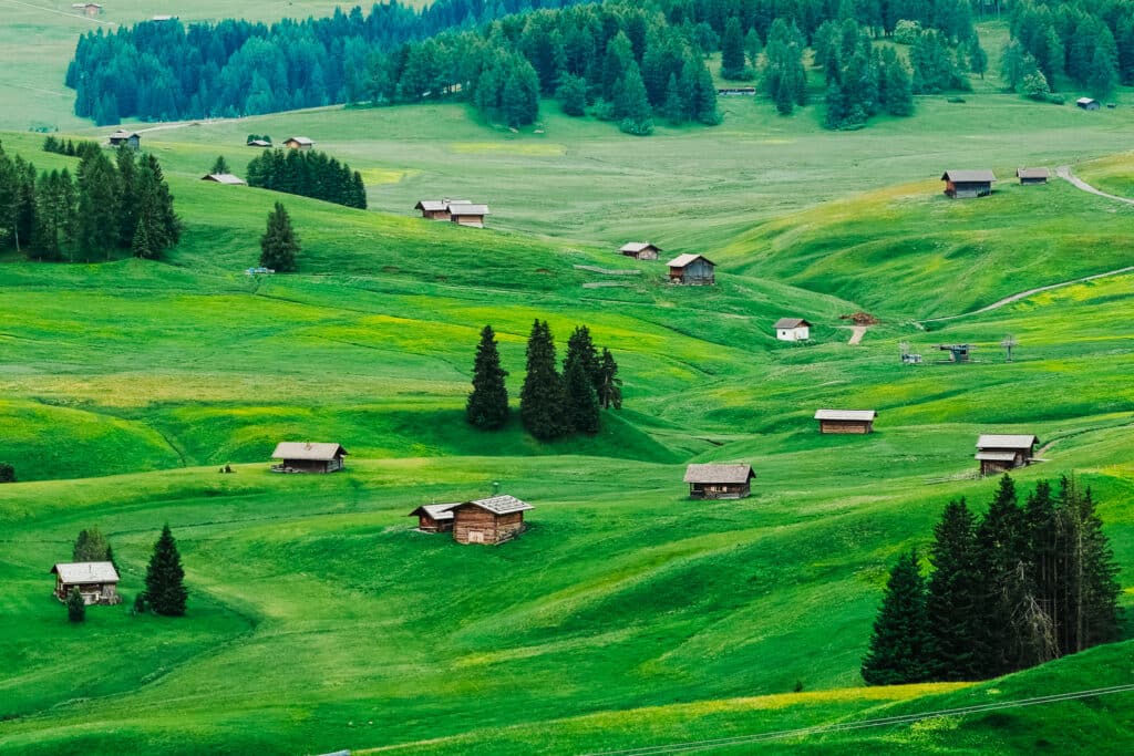 Aerial view of Alpe di Siusi plateau in the Dolomites, South Tyrol, with rolling emerald green meadows and traditional wooden alpine huts dotted across the hills surrounded by pine trees