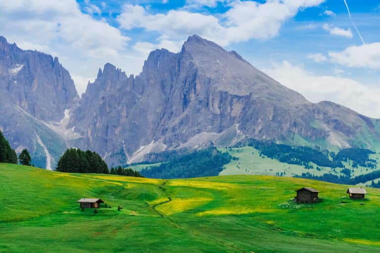 Iconic view of Sassolungo and Sassopiatto peaks towering over bright green meadows dotted with traditional wooden mountain huts at Alpe di Siusi, Dolomites, in summer