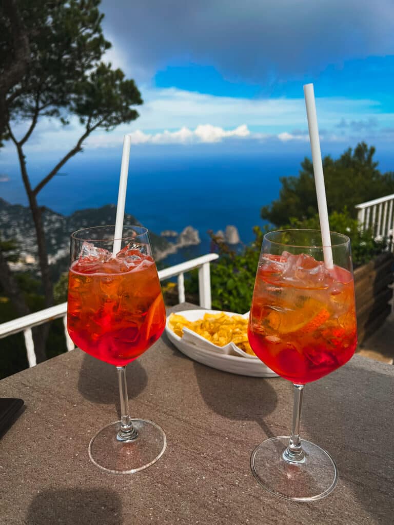 Two Aperol Spritz cocktails on a terrace table with a stunning view of the Faraglioni rocks and Capri coastline