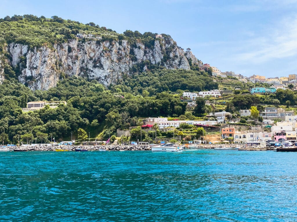 Capri's marina viewed from the water, with turquoise sea, moored boats, and white buildings climbing the rocky green cliffs