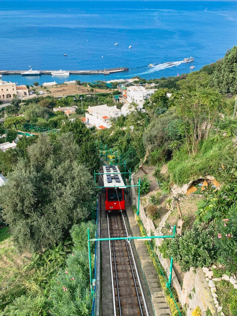 Capri's red funicular descending through lush terraced gardens toward the marina, with the blue sea visible below