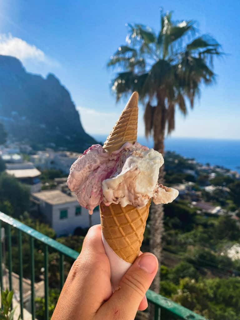 Hand holding a double-scoop gelato cone with Capri town, a palm tree, and mountain cliffs in the sunny background