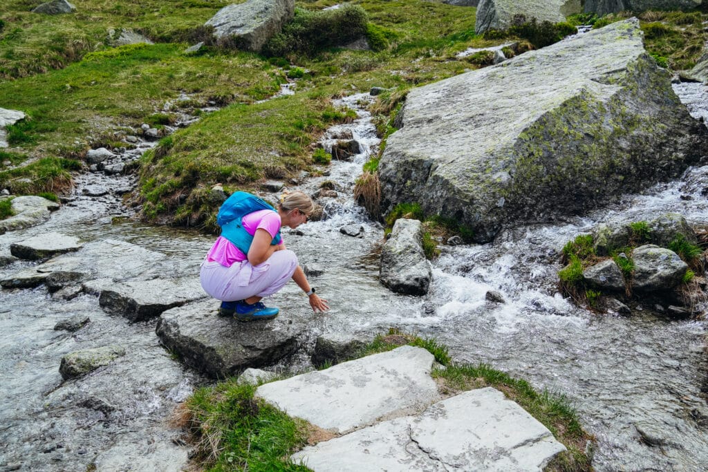 Hiker crouching to dip her hand in a clear alpine stream cascading over granite slabs on the Grand Balcon Nord