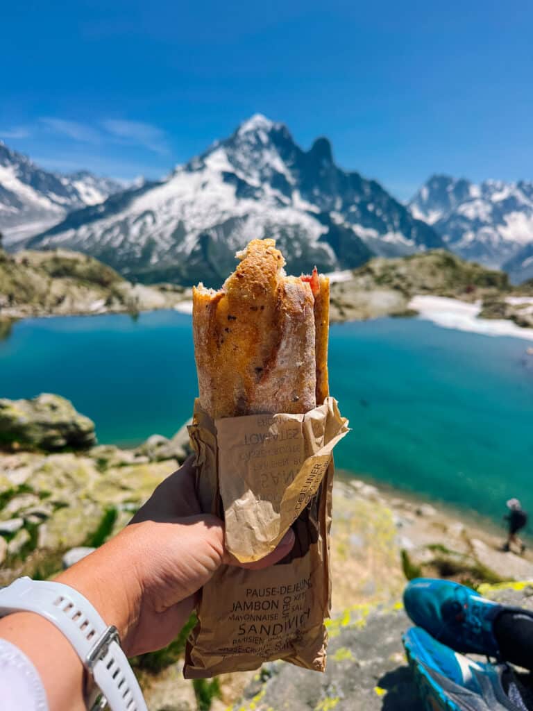 Hand holding a French jambon baguette sandwich above the bright blue water of Lac Blanc with snow-capped peaks behind