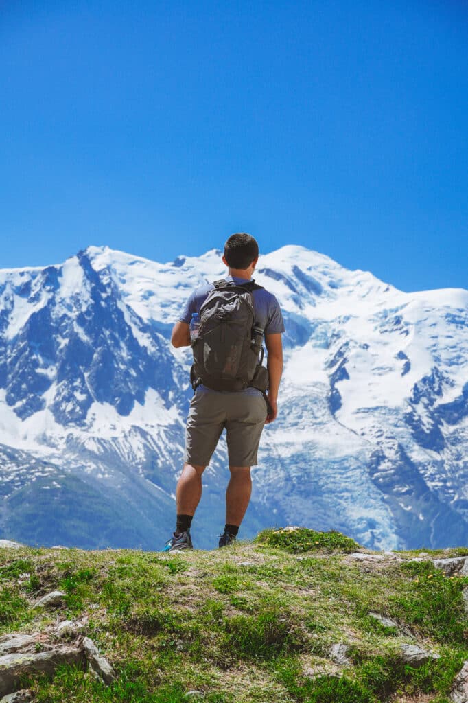 Male hiker with a backpack standing on a grassy ridge admiring the snow-covered Mont Blanc range above Chamonix valley