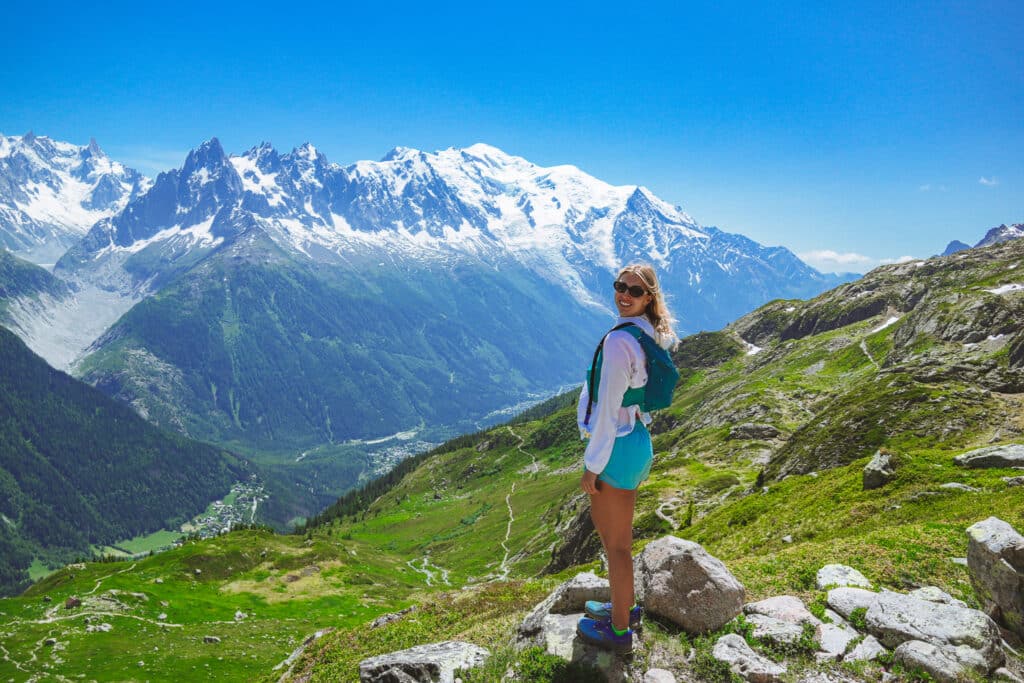 Female hiker in turquoise shorts looking back toward the camera with the full Mont Blanc massif spread behind her