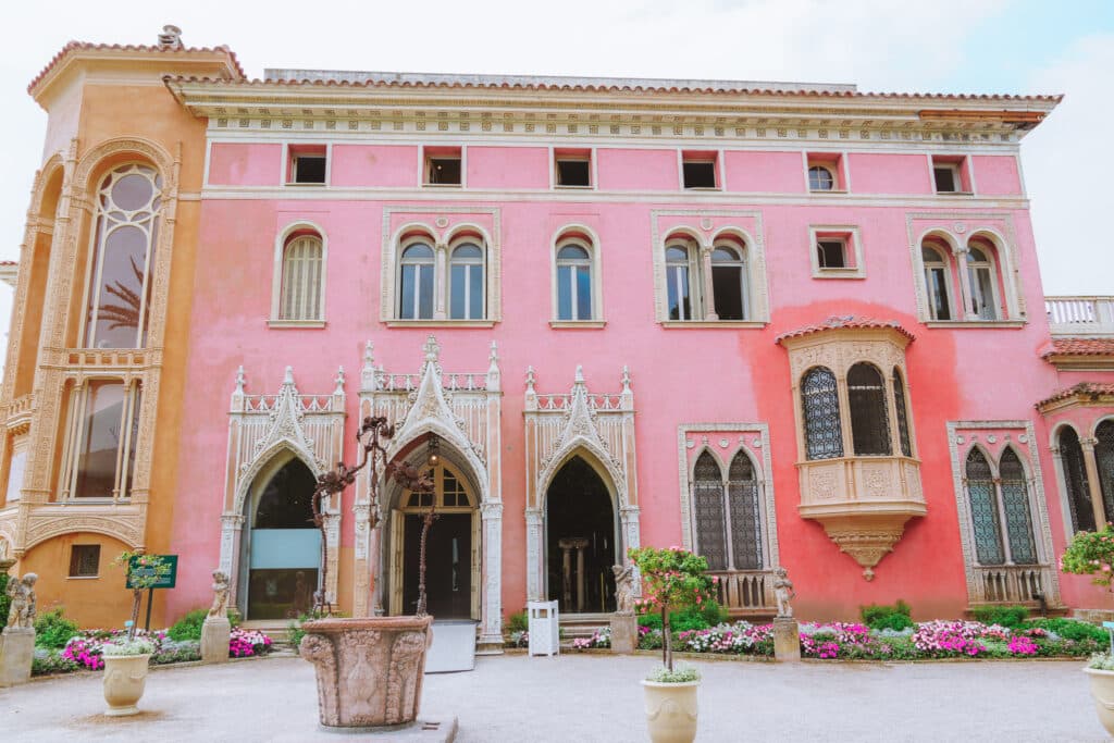 The pink facade of the Villa Ephrussi de Rothschild with a white pebbled driveway, and planters with little citrus trees, and a wishing well