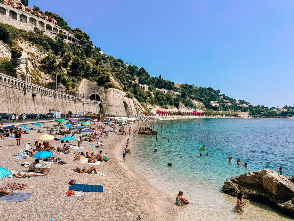 A bright sunny day at Plage des Marinieres in Villefranche-sur-Mer with beach goers sunning themselves towel to towel on the pebbled shores