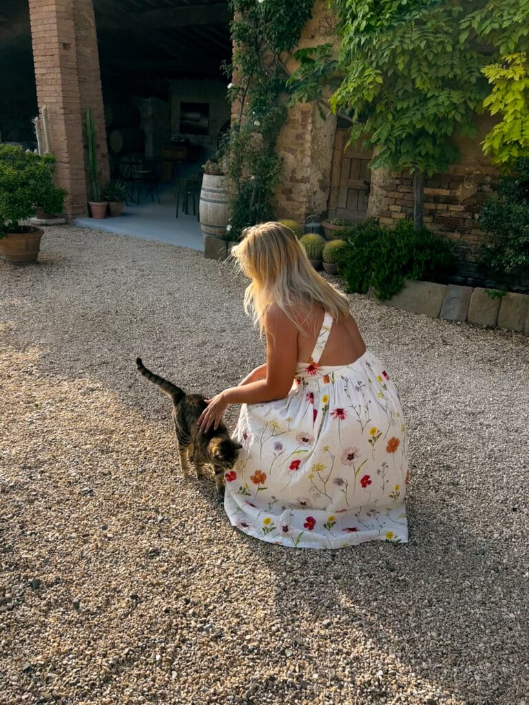 Guest in a floral dress crouching to greet a tabby cat in the gravel courtyard at Agriturismo Marciano in Tuscany.
