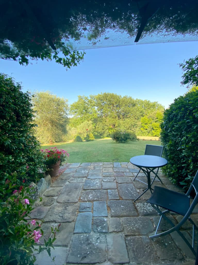 View from the guesthouse patio at Agriturismo Marciano, looking past potted geraniums onto a sunny garden lawn in Tuscany.