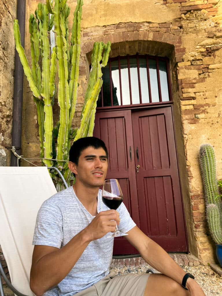 Traveler relaxing in a deck chair with a glass of red wine beside tall cacti and a burgundy door at Agriturismo Marciano.
