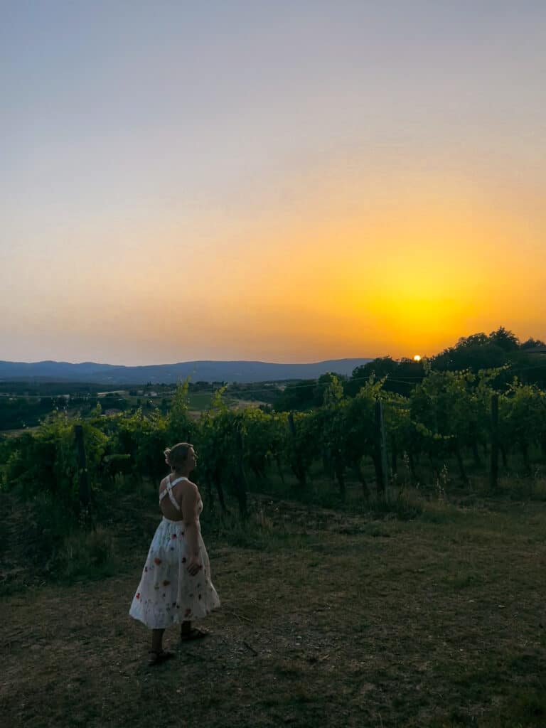Woman in a floral dress watching a golden sunset over the vineyard rows at Agriturismo Marciano in Tuscany.