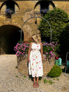 Woman in a floral summer dress posing in front of the geranium-covered stone well in the courtyard at Agriturismo Marciano, Tuscany.