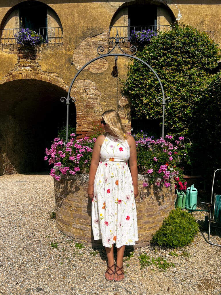 Woman in a floral summer dress posing in front of the geranium-covered stone well in the courtyard at Agriturismo Marciano, Tuscany.